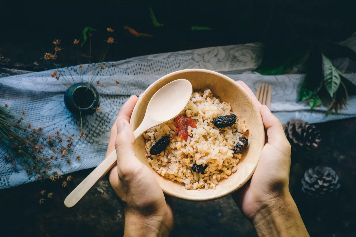 Hands holding a wooden bowl of rice with dried fruits and a wooden spoon