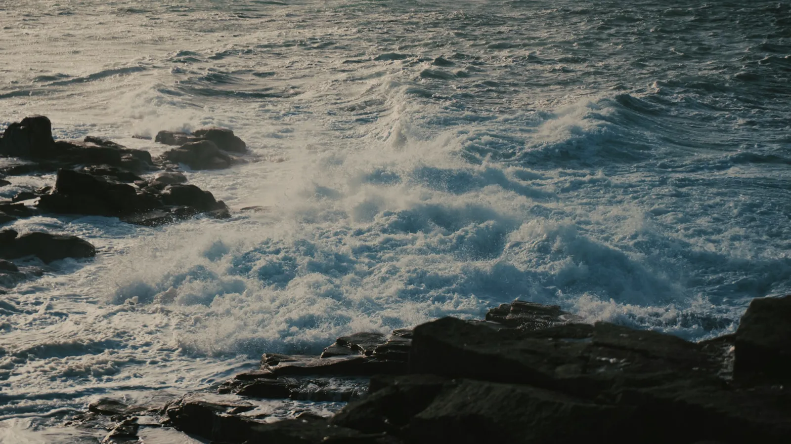 Ocean waves crash against dark volcanic rocks along a rugged coastline at golden hour, the white foam contrasting with the deep blue sea — evoking the natural, individual rhythm of moving water finding its own way.