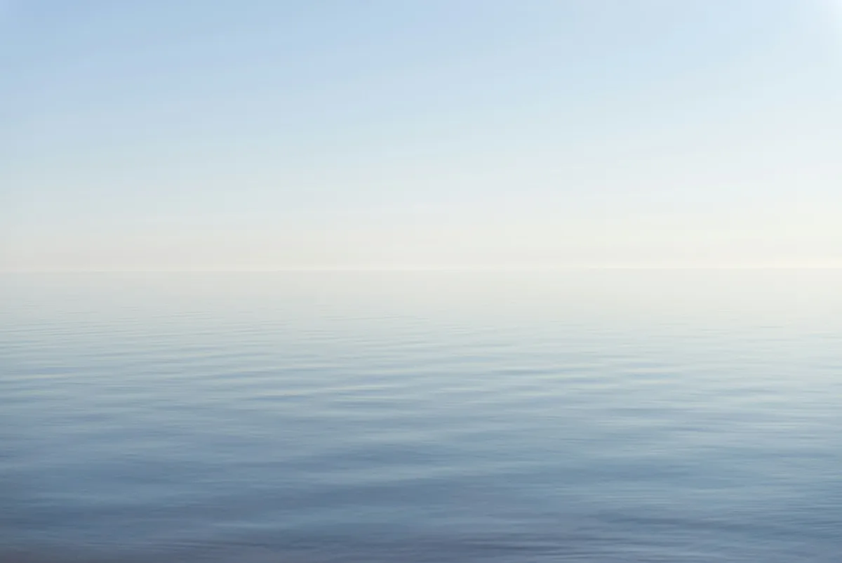 A calm sea meeting a pale sky at the horizon, with gentle ripples softening the surface