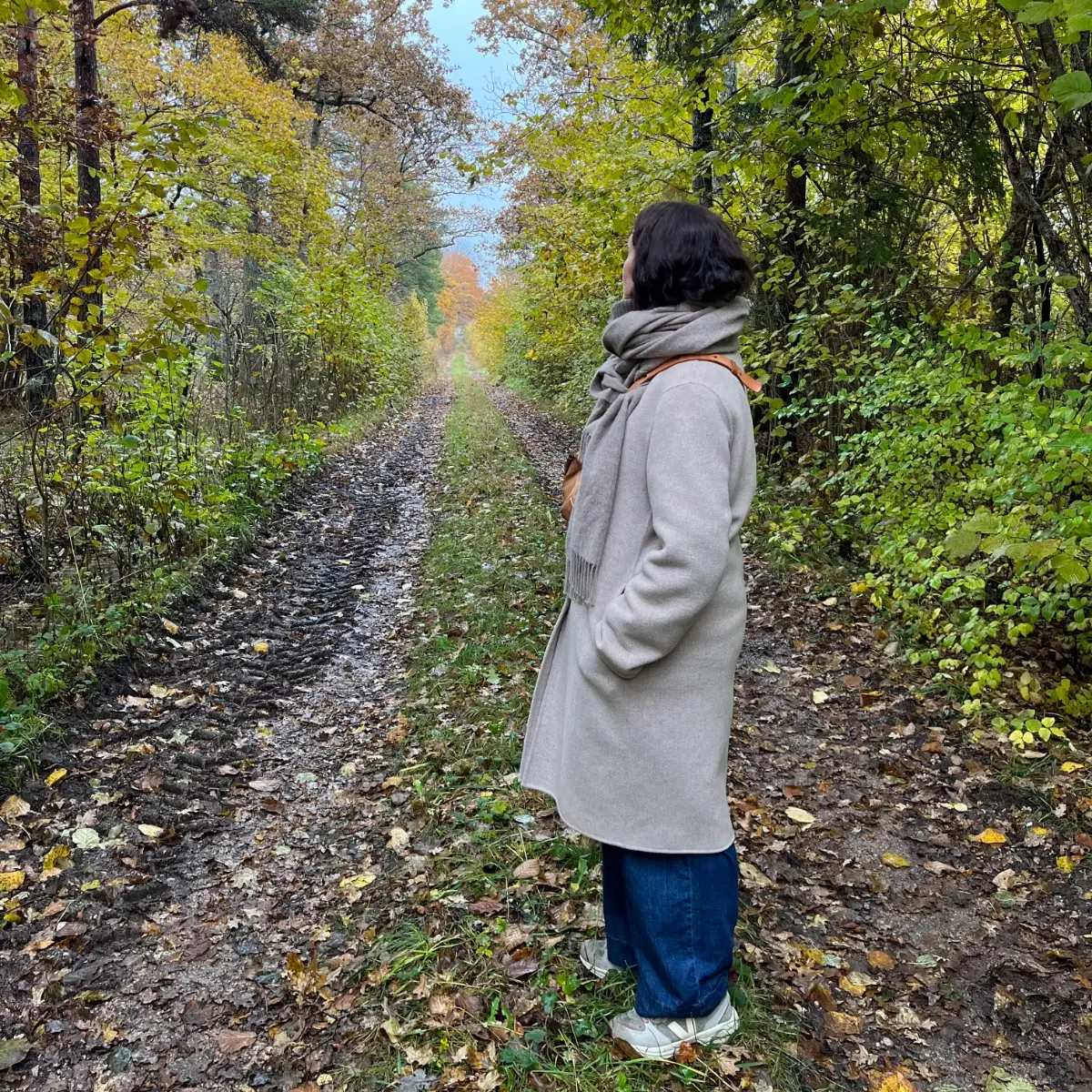 A person wearing a beige coat and scarf stands on a muddy forest path surrounded by lush green and autumn-colored trees, looking into the distance. The scenic woodland trail features fallen leaves, tire tracks, and a peaceful natural atmosphere, ideal for hiking, mindfulness, and outdoor exploration during fall.
